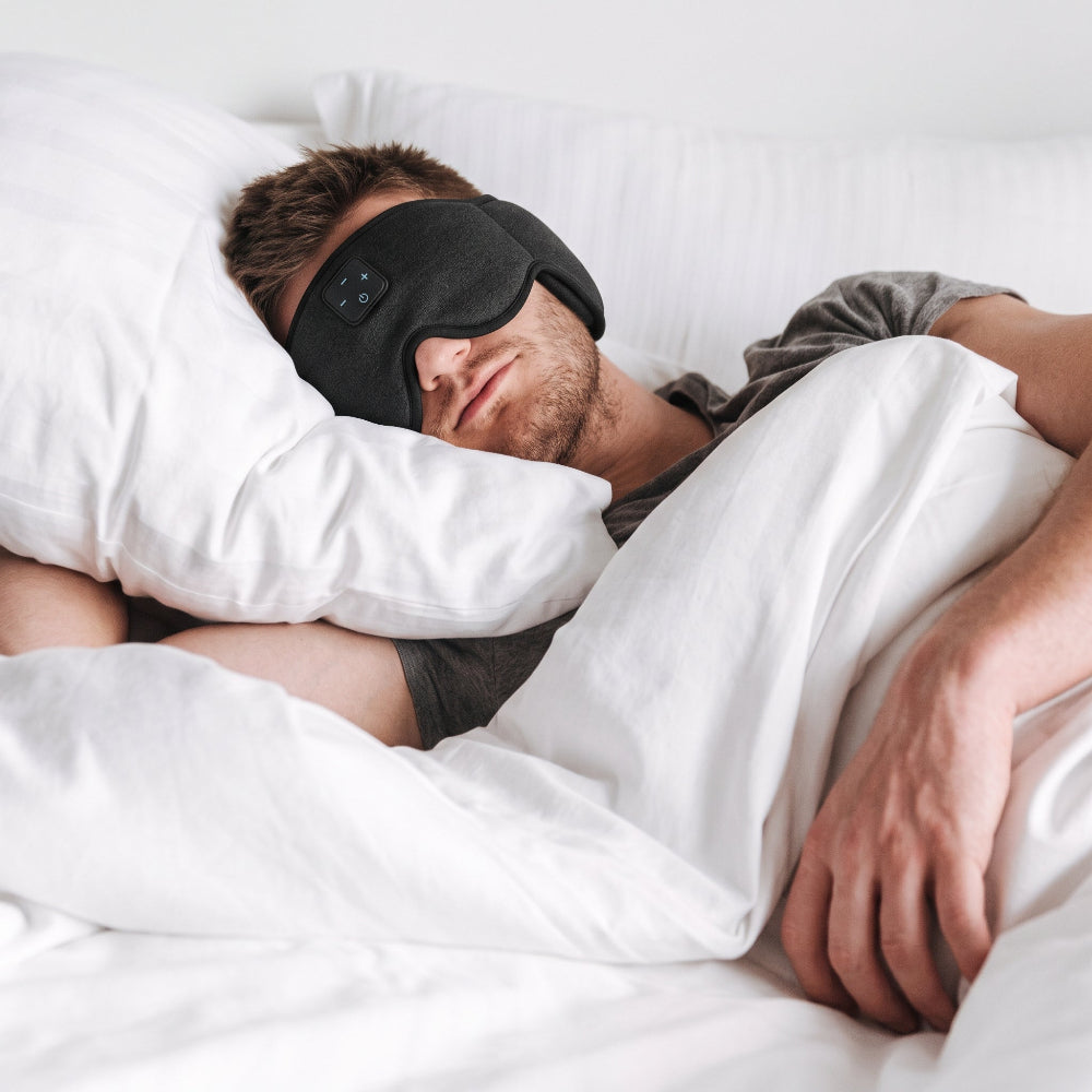 A man lying in bed under white sheets, wearing a black sleep mask with a small control panel on the side.